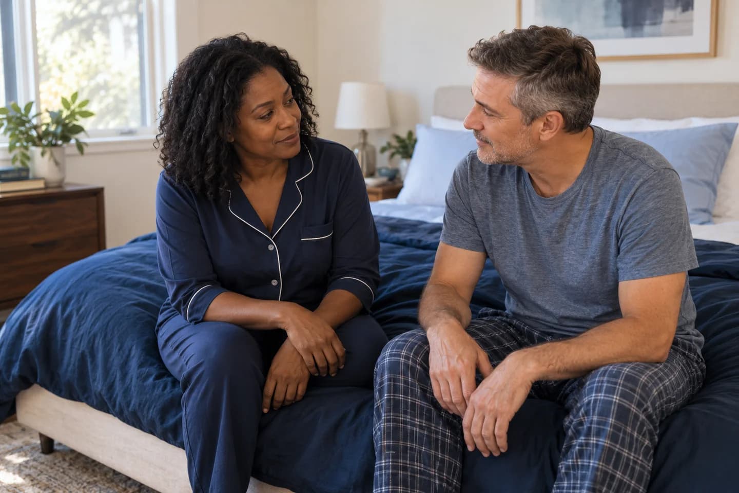 Two adults sit on the edge of a bed and talk in soft morning light.